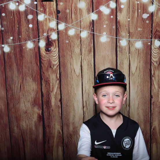Ein junger Junge mit schwarzer Kappe und Weste steht vor einem hölzernen Hintergrund mit Lichtern. Er lächelt für die Kamera.