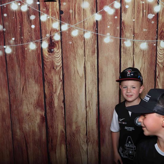 Zwei Jungen posieren für ein Foto vor einem hölzernen Hintergrund mit Lichterketten, einer trägt einen Baseballcap und der andere einen schwarzen Hut.