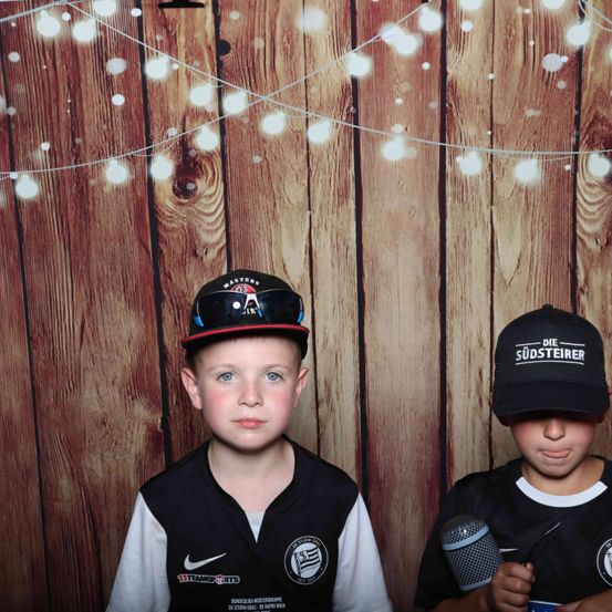 Zwei junge Jungen posieren für ein Foto vor einem hölzernen Hintergrund mit Lichtern. Ein Junge trägt einen schwarzen Hut mit 'Masters' und eine Weste mit Logo, während der andere ein Mikrofon hält und einen schwarzen 'Die Sudsteirer' Hut trägt.