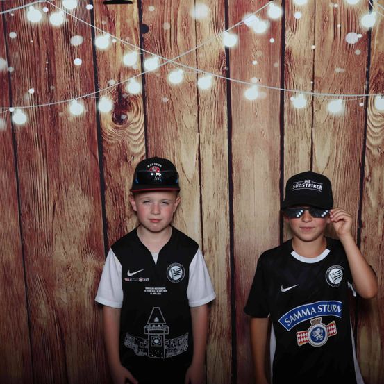 Zwei Jungen in Sporttrikots und Mützen stehen vor einer Holzwand mit Lichterketten. Der Junge auf der linken Seite trägt ein schwarzes Weste mit Nike-Logo und einem Uhrturm-Design. Der Junge auf der rechten Seite trägt ein schwarzes Trikot mit dem Text 'SAMMA STURM' und Sonnenbrille.