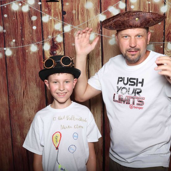 Ein Mann und ein Junge posieren für ein Foto. Der Mann trägt einen braunen Hut und ein weißes T-Shirt mit der Aufschrift 'PUSH YOUR LIMITS'. Der Junge trägt einen schwarzen Hut mit Schutzbrille und ein weißes T-Shirt mit Zeichnungen.