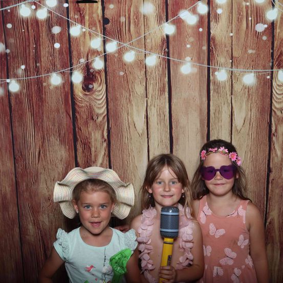 Drei junge Mädchen lächeln für ein Foto in einer Fotokabine. Eine hält ein Mikrofon, eine andere trägt eine Blumenkrone. Der Hintergrund ist eine Holzwand mit Lichterketten.