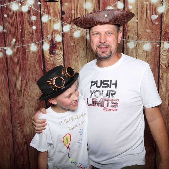 Ein Mann mit einem Piratenhut und einem weißen T-Shirt mit den Worten 'PUSH YOUR LIMITS' steht neben einem Jungen, der einen schwarzen Hut und eine Schutzbrille trägt. Sie lächeln beide in die Kamera. Der Hintergrund ist eine Holzwand mit Lichtern.