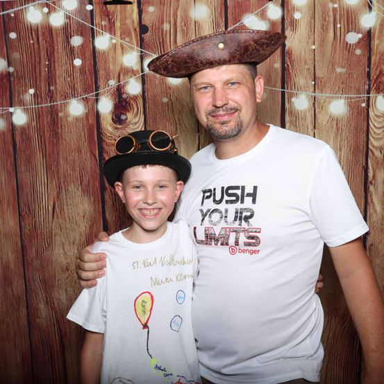 Ein Mann und ein Junge stehen nah beieinander und lächeln für ein Foto. Der Mann trägt einen Piratenhut und ein weißes T-Shirt mit 'PUSH YOUR LIMITS'. Der Junge trägt einen Hut, eine Brille und ein weißes T-Shirt mit einem Ballon-Zeichnung. Sie posieren vor einem hölzernen Hintergrund mit Lichterketten.