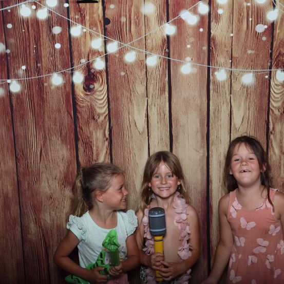 Drei junge Mädchen stehen vor einer Holzwand mit Lichterketten. Sie lächeln und halten Spielzeug. Ein Mädchen hält ein Mikrofon.