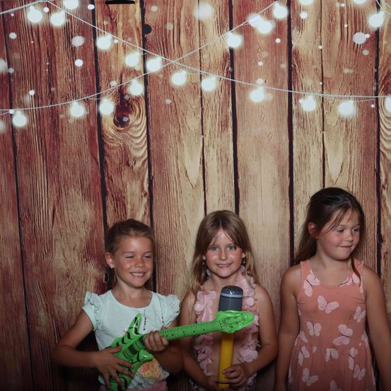 Drei junge Mädchen posieren gemeinsam vor einem hölzernen Hintergrund mit Lichterketten. Eine hält eine Spielzeuggitarre, eine andere ein Spielzeugmikrofon und die dritte lächelt direkt in die Kamera.