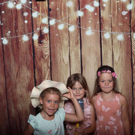 Drei junge Mädchen posieren vor einem hölzernen Hintergrund, der mit Lichterketten geschmückt ist. Ein Mädchen trägt einen Strohhut, ein anderes hält ein Mikrofon und das dritte ein Schmetterlingskleid.