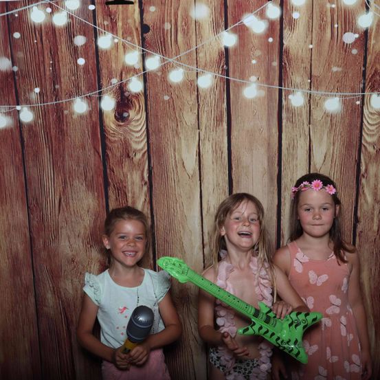Drei junge Mädchen stehen vor einem hölzernen Hintergrund, jede hält ein Requisit. Das Mädchen auf der linken Seite hält ein Mikrofon, die in der Mitte eine grüne Gitarre und das Mädchen rechts eine Blumenkrone. Über ihnen sind Lichterketten befestigt.