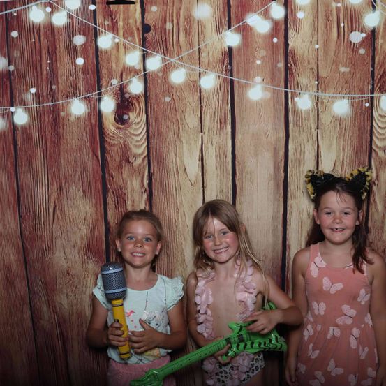 Drei junge Mädchen stehen vor einer Holzwand. Das Mädchen links hält ein Mikrofon, das Mädchen in der Mitte eine Spielzeuggitarre und das Mädchen rechts trägt ein Schmetterlingskleid. Sie alle lächeln.