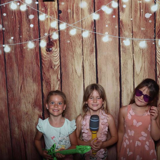 Drei junge Mädchen stehen vor einem mit Lichterketten verzierten Holzhintergrund. Das Mädchen links hält eine Spielzeuggitarre, die in der Mitte hat ein Mikrofon und das rechte trägt eine Sonnenbrille.