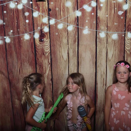 Drei junge Mädchen stehen vor einer mit Lichterketten geschmückten Holzwand, eine hält eine Spielzeuggitarre und eine andere ein Mikrofon. Die Mädchen scheinen Spaß zu haben, wahrscheinlich in einem Fotokabine.