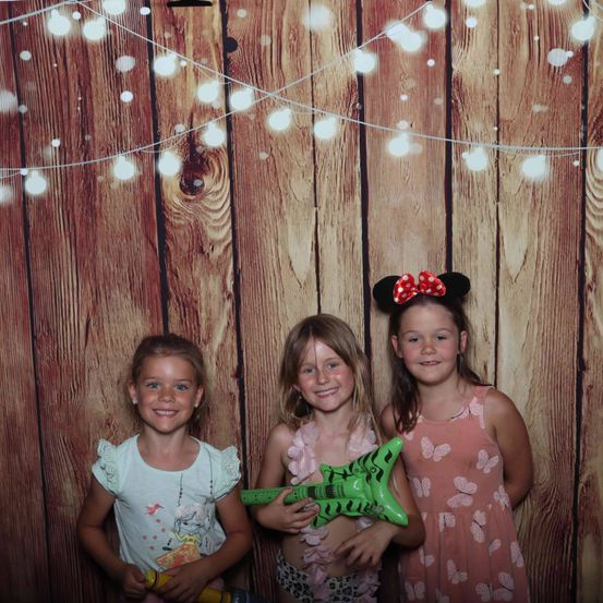 Drei junge Mädchen stehen vor einer Holzwand mit Lichtern, tragen Sommerkleider und lächeln. Das Mädchen in der Mitte hält eine grüne Spielzeuggitarre.