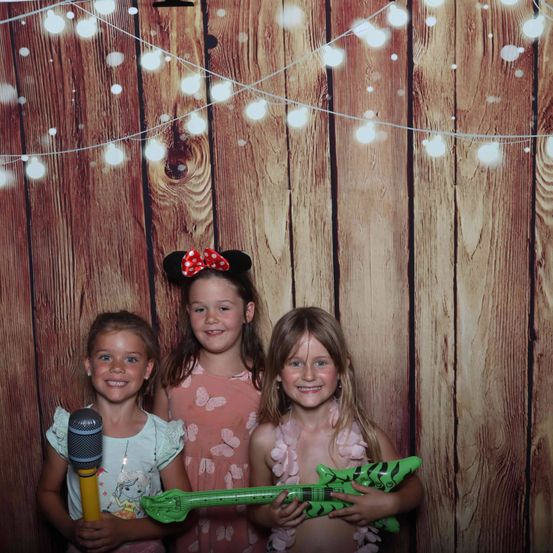 Drei junge Mädchen posieren vor einem hölzernen Hintergrund mit Lichterketten und halten ein Mikrofon und eine grüne Spielzeuggitarre. Sie lächeln alle.