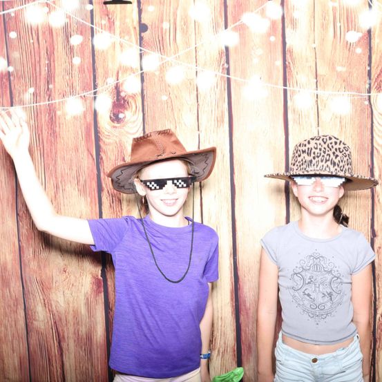 Zwei junge Mädchen stehen gemeinsam vor einer Holzwand mit Lichtern. Sie tragen beide Cowboyhüte und Sonnenbrillen. Das Mädchen auf der linken Seite trägt ein lila T-Shirt und das Mädchen auf der rechten Seite ein graues T-Shirt.