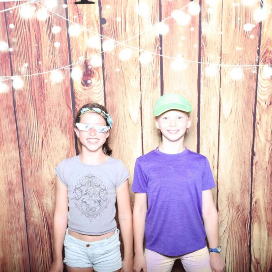 Zwei Kinder stehen vor einer Holzwand, ein Mädchen mit einem grauen Shirt und Shorts und ein Junge mit einem lila Shirt, beide lächeln.