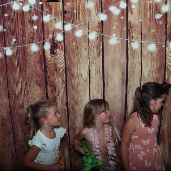 Drei junge Mädchen posieren vor einem hölzernen Hintergrund mit Lichterketten. Das linke Mädchen schaut nach oben, das mittlere Mädchen hält eine grüne Puppe und das rechte Mädchen schaut zur Seite.