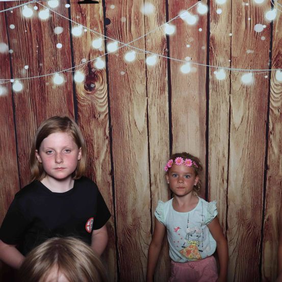 Zwei junge Mädchen posieren vor einem hölzernen Hintergrund mit Lichterketten. Ein Mädchen trägt eine rosa Haarschleife und ein weißes Oberteil, während das andere ein schwarzes T-Shirt trägt.