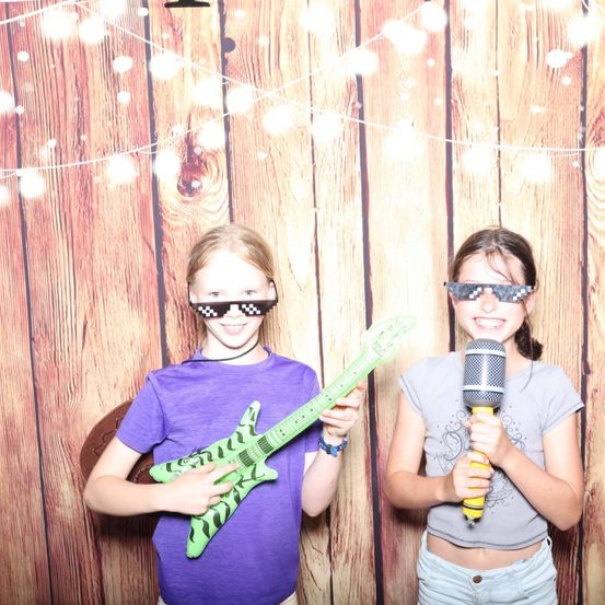 Zwei Mädchen posieren mit Requisiten. Eines hält eine grüne Spielzeuggitarre, das andere ein Mikrofon. Beide tragen Brillen. Ein Holzwand mit Lichterketten steht dahinter.