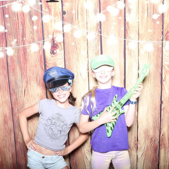 Zwei junge Mädchen stehen nebeneinander, eine hält eine grüne Gitarre und beide tragen Hüte und Brillen. Sie lächeln und scheinen für ein Foto zu posieren. Der Hintergrund ist eine Holzwand mit Lichterketten.