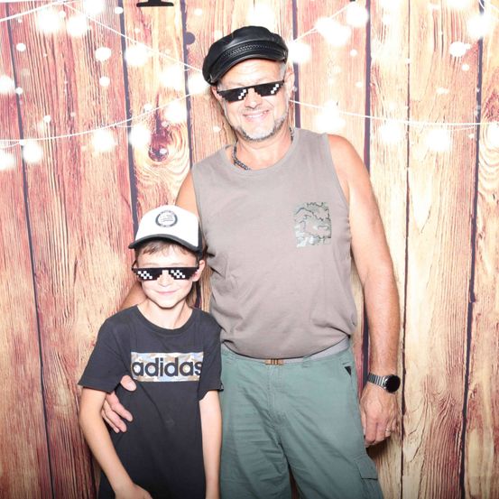 Ein erwachsener Mann mit Sonnenbrille und Hut steht mit einem jungen Jungen, der eine Baseballkappe und Sonnenbrille trägt, beide lächeln. Sie stehen vor einer Holzwand mit dekorativen Lichtern.