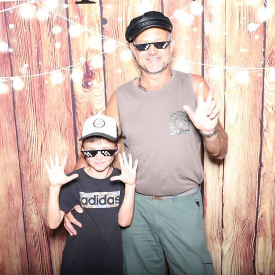 Ein Mann und ein Junge, beide mit Sonnenbrille, stehen eng beieinander und posieren für ein Foto. Der Mann trägt eine Mütze und eine Halskette, der Junge eine Kappe. Sie machen Friedenszeichen. Hinter ihnen ist ein hölzerner Hintergrund.