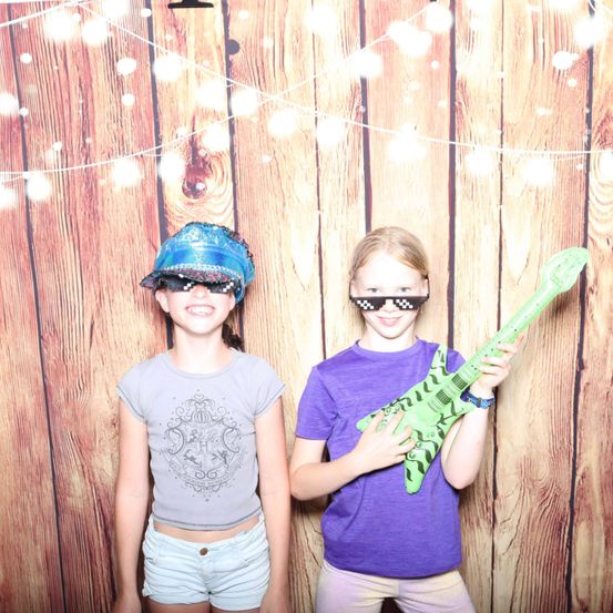 Zwei junge Mädchen in einem Fotostudio, die eine Sonnenbrille tragen und eine grüne Gitarre halten. Das Mädchen auf der linken Seite trägt einen Hut und ein T-Shirt mit einem grafischen Design. Das Mädchen auf der rechten Seite trägt ein lila T-Shirt und lächelt. Der Hintergrund ist eine Holzwand mit weißen Lichterketten.
