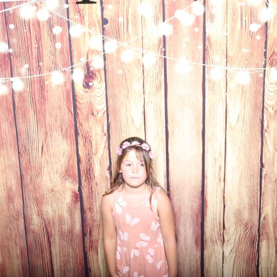 Ein junges Mädchen steht vor einem hölzernen Hintergrund mit Schmetterlingsdekorationen und Lichterketten. Sie trägt ein rosa Kleid und eine Blumenkrone.