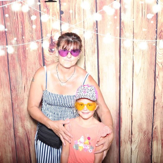 Eine erwachsene Frau und ein junges Mädchen posieren vor einem hölzernen Hintergrund. Die Frau trägt ein gestreiftes Tanktop und lila Sonnenbrille, während das Mädchen eine Mütze und gelbe Brille trägt. Beide lächeln.
