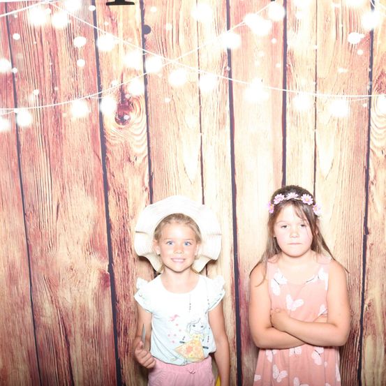 Zwei junge Mädchen posieren vor einem hölzernen Hintergrund mit dekorativen Lichtern. Eine trägt einen weißen Hut, die andere eine Blumenkrone.