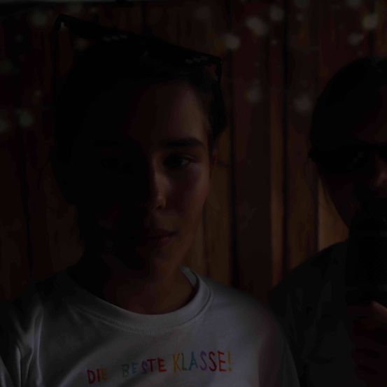 Eine junge Frau mit einem weißen T-Shirt, auf dem 'DIE BESTE KLASSE!' steht, lächelt in einem schwach beleuchteten Raum. Daneben steht eine weitere Person mit einem Telefon.