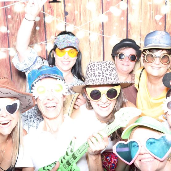 Eine Gruppe lächelnder Frauen mit verschiedenen Hüten und Sonnenbrillen posiert für ein Foto in einem Studio mit hölzerne Kulisse.