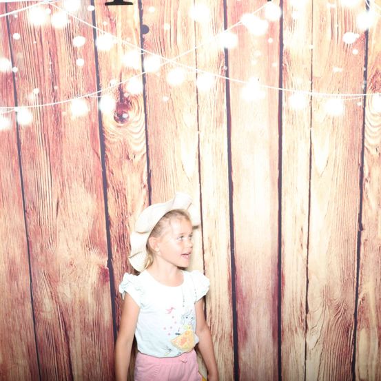 Ein kleines Mädchen in einem weißen Hut und rosa Hosen steht vor einer Holzwand mit Lichterketten.