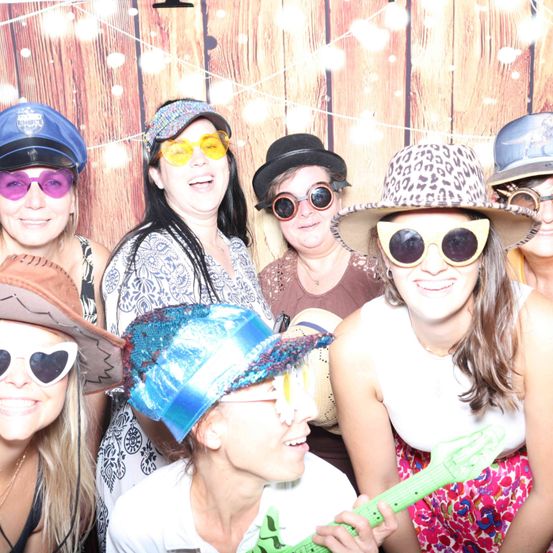 Eine Gruppe von Frauen in Hüten und Sonnenbrillen posiert für ein Foto vor einem hölzernen Hintergrund mit Lichtern.