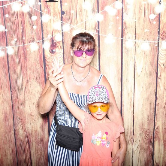 Eine Frau mit Sonnenbrille und ein junges Mädchen mit Hut und Brille stehen zusammen vor einem hölzernen Hintergrund.