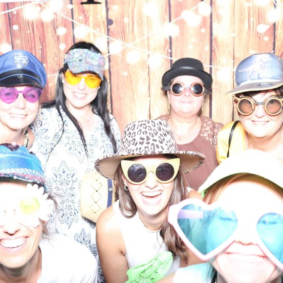 Eine Gruppe von Frauen, die Hüte und Sonnenbrillen tragen, lächeln in einem Fotokabine mit hölzerne Hintergrund.