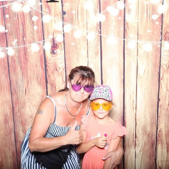 Eine Frau und ein junges Mädchen stehen vor einem hölzernen Hintergrund. Die Frau zeigt Daumen hoch, während das Mädchen einen Lolli hält. Beide tragen Sonnenbrillen.