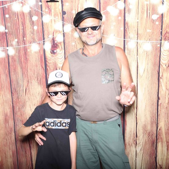 Ein Mann und ein Junge stehen vor einer Holzwand. Der Mann trägt eine Sonnenbrille und einen Hut. Der Junge trägt einen Hut und ein schwarzes T-Shirt mit weißen Text.