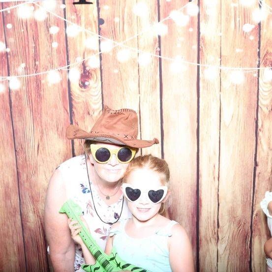 Eine Frau und ein junges Mädchen lächeln für ein Foto. Sie tragen Sonnenbrillen und einen Cowboyhut. Das Mädchen hält eine grüne Gitarre. Hinter ihnen ist eine Holzwand mit Lichterketten.