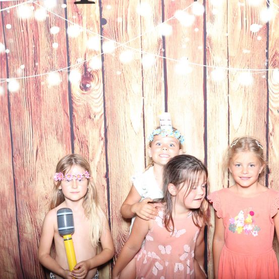 Vier junge Mädchen posieren vor einem mit Lichtern geschmückten hölzernen Hintergrund. Ein Mädchen hält ein Mikrofon, während die anderen Blumenkränze und Kleider tragen.