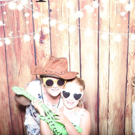 Eine Frau und ein junges Mädchen posieren für ein Foto in einem Fotokabine. Die Frau trägt einen Cowboyhut und eine Sonnenbrille. Das Mädchen hält eine grüne Gitarre und trägt eine Herzform-Sonnenbrille. Sie lächeln und stehen vor einer Holzwand mit Lichtern.