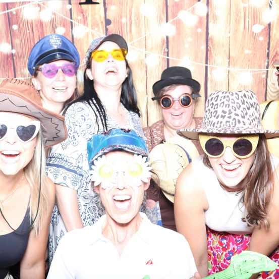 Eine Gruppe von Menschen posiert für ein Foto vor einem hölzernen Hintergrund. Sie tragen verschiedene Hüte und Sonnenbrillen, eine Person hält eine grüne Gitarre.