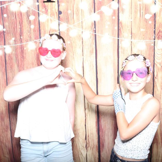 Zwei junge Mädchen stehen vor einem hölzernen Hintergrund und tragen herzförmige Sonnenbrillen und Blumenkränze. Sie lächeln und eine hält ihre Hände in Herzform.