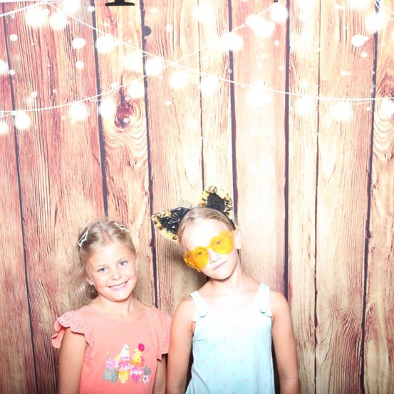 Zwei junge Mädchen stehen vor einem hölzernen Hintergrund, eines trägt orangefarbene Brille und eine Schleife im Haar, das andere ein rosa Shirt. Sie lächeln fröhlich.