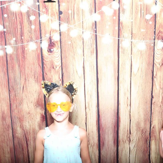 Ein junges Mädchen in einem blauen Kleid und gelben herzförmigen Sonnenbrillen posiert vor einer Holzwand mit Lichterketten.