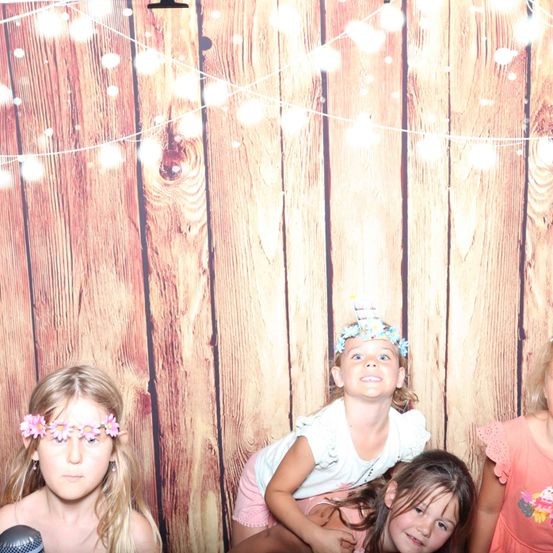 Drei junge Mädchen stehen zusammen in einer Fotokabine. Eine trägt eine Blumenkrone, eine andere ein kuchenthematisches Kleid und die dritte hält ein Mikrofon. Der Hintergrund besteht aus Holzpaneelen und dekorativen Lichtern.