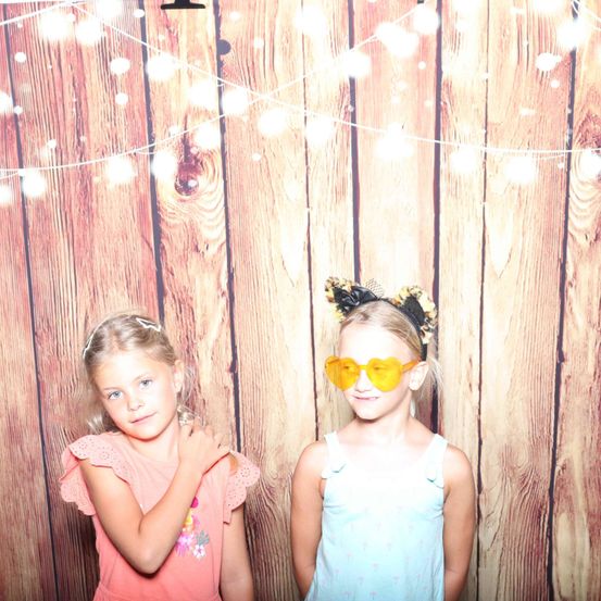 Zwei junge Mädchen stehen vor einer Holzwand. Sie tragen Kleider und haben die Arme verschränkt. Das Mädchen rechts trägt eine Sonnenbrille. Die Wand hat Lichterketten.