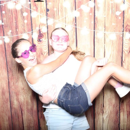 Zwei Mädchen lächeln und posieren für ein Foto in einem Fotoautomaten. Sie tragen Sonnenbrillen und eine hält die Beine der anderen.