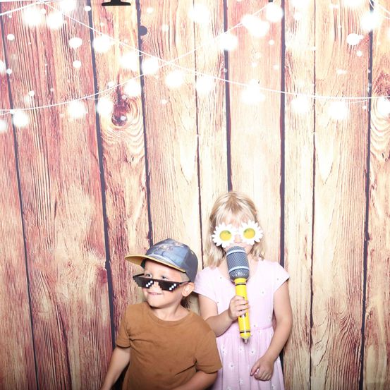 Zwei Kinder stehen vor einem mit Lichterketten geschmückten Holzhintergrund. Ein Junge trägt eine Mütze und eine Sonnenbrille, während ein Mädchen ein Mikrofon hält und blumenförmige Brillen trägt.