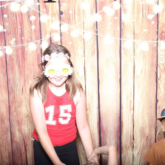 Ein Mädchen mit Blumenkopfband und weißen Gläsern posiert für ein Foto vor einem hölzernen Hintergrund mit Lichterketten.