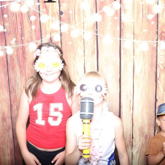 Zwei Mädchen posieren in einem Fotokabine. Eines hält ein Mikrofon, und die andere trägt ein rotes Trikot mit der Nummer 15. Dahinter ist eine Holzwand mit Lichtern.
